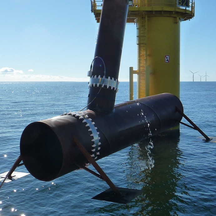 An einer Offshore-Gründungsstruktur  ist eine Sensormanschette zur Überwachung von Schweißnähten befestigt. Im Hintergrund sieht man das Meer, den blauen Himmel und Windräder.